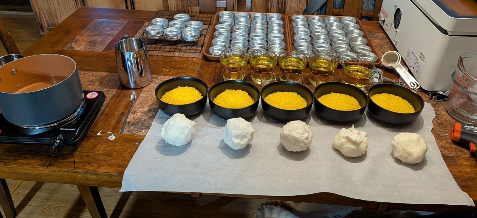 Batch day on the kitchen table: bowls of beeswax, balls of shea butter, and measured oils lined up beside trays of empty tins